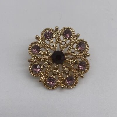 Broche vintage en métal doré (ou ton or) ornée de strass violets (type améthyste) et de cristaux clairs, ressemblant à un motif floral ou à une rosace