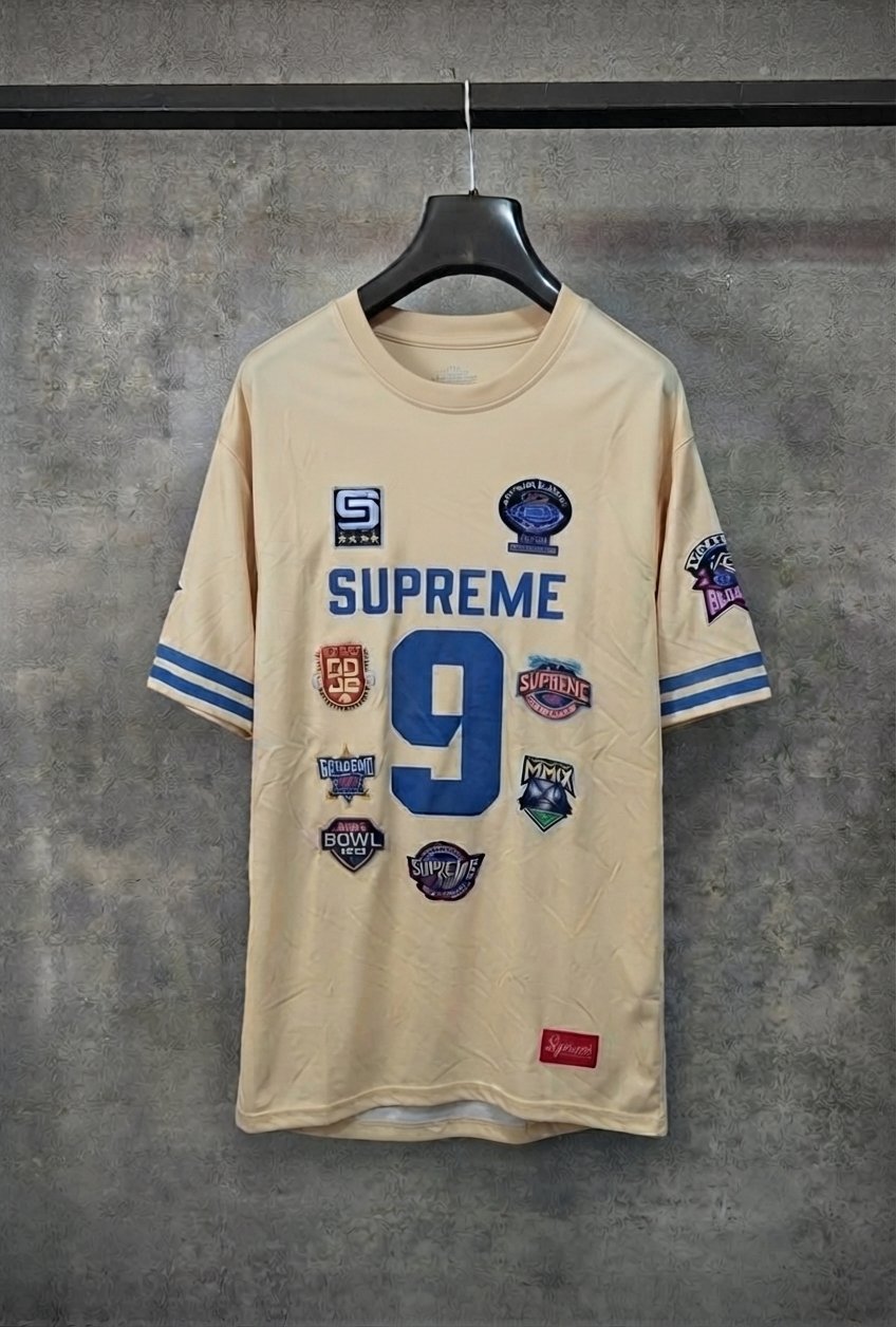 Ce maillot est le Supreme Championships Embroidered Football Jersey de la collection Automne/Hiver 2023, reconnaissable à son coloris doré et ses multiples écussons brodés