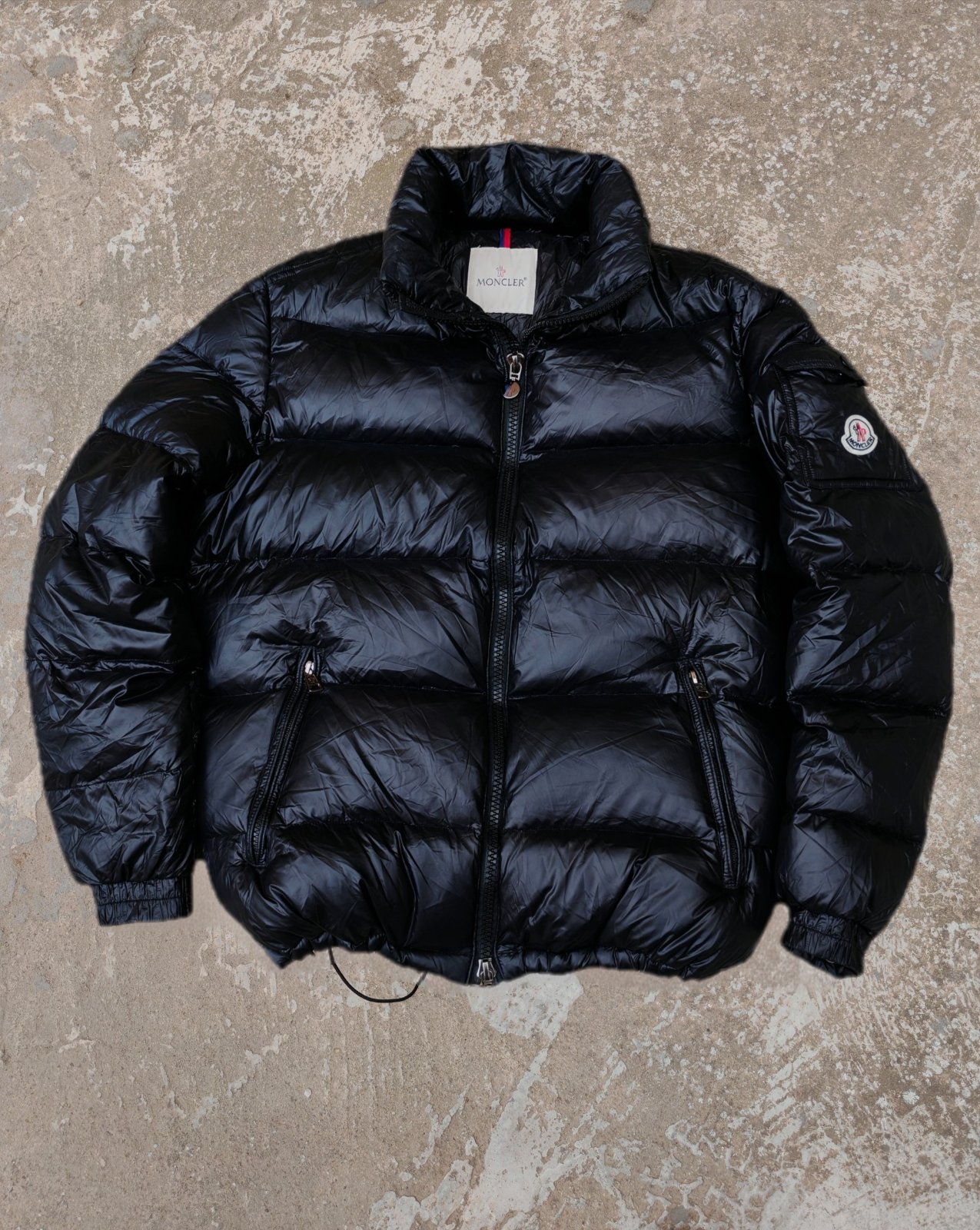 Cette pièce est le modèle emblématique Moncler Maya, une doudoune courte pour homme en nylon laqué brillant noir caractérisée par ses boudins de matelassage horizontaux et sa poche plaquée signature sur le bras gauche arborant le logo de la marque.