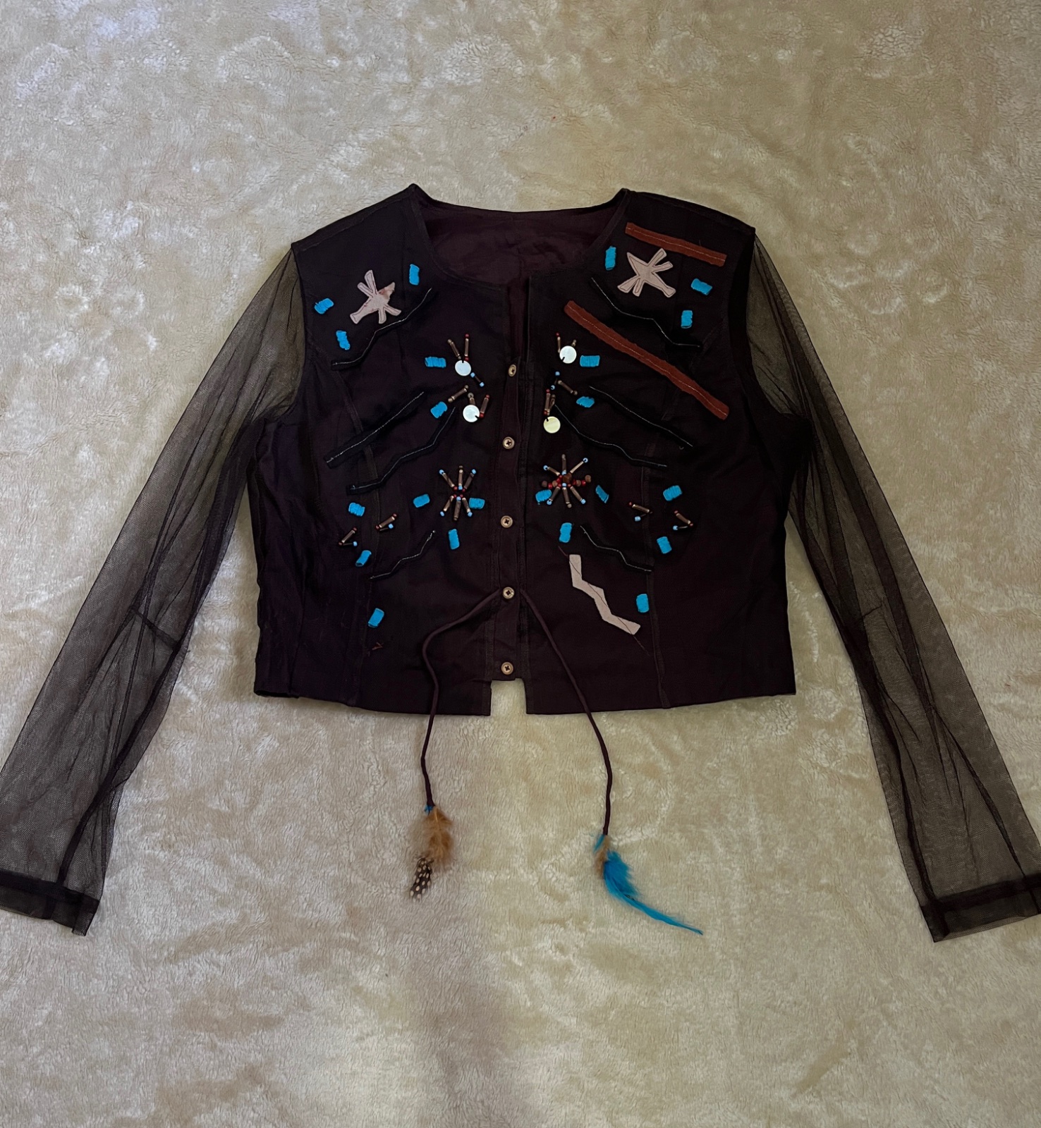 Brown boho-style vest featuring sheer mesh long sleeves, intricate beadwork, turquoise accents, and decorative feathers
