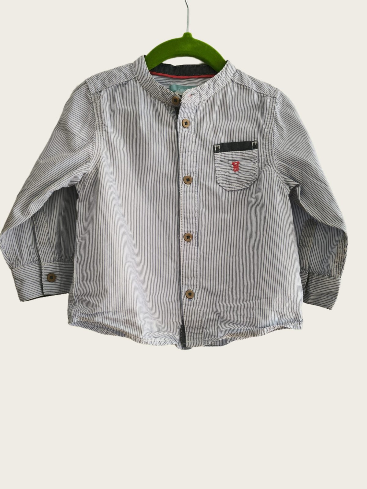Une chemise à manches longues pour bébé garçon à rayures verticales bleu et blanc, dotée d'un col mao et d'une poche poitrine contrastée, qui correspond au modèle de la marque Obaïbi.