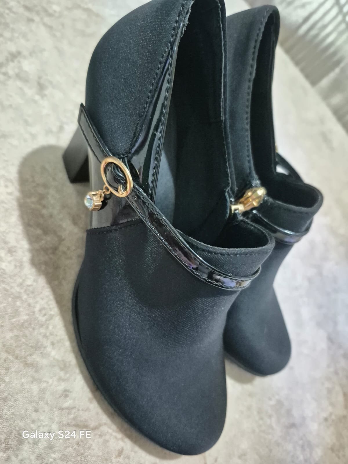 Une paire de bottines élégantes pour femme en suédine noire (ou daim), caractérisée par une découpe basse, un talon épais et une bride décorative en similicuir verni sur le dessus, agrémentée d'une boucle dorée et d'un petit pendentif brillant.