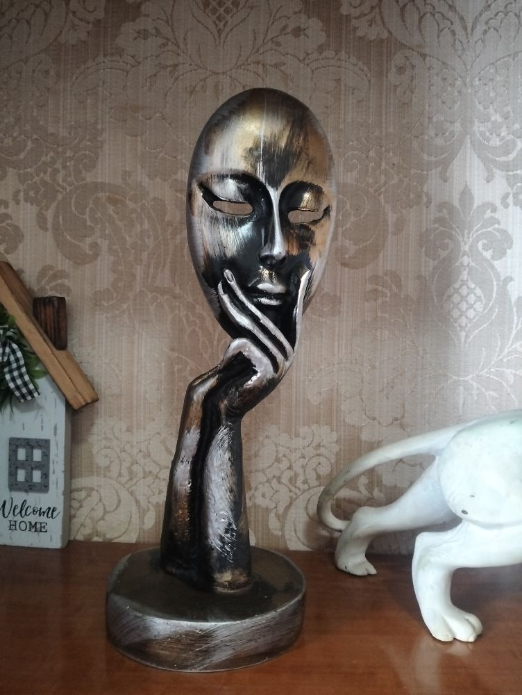 Sculpture décorative représentant un visage féminin de type masque en pleine contemplation ,tenu par une main allongée et stylisée ,avec une finition métalisée principalement de couleur argentée avec des accents de bronze