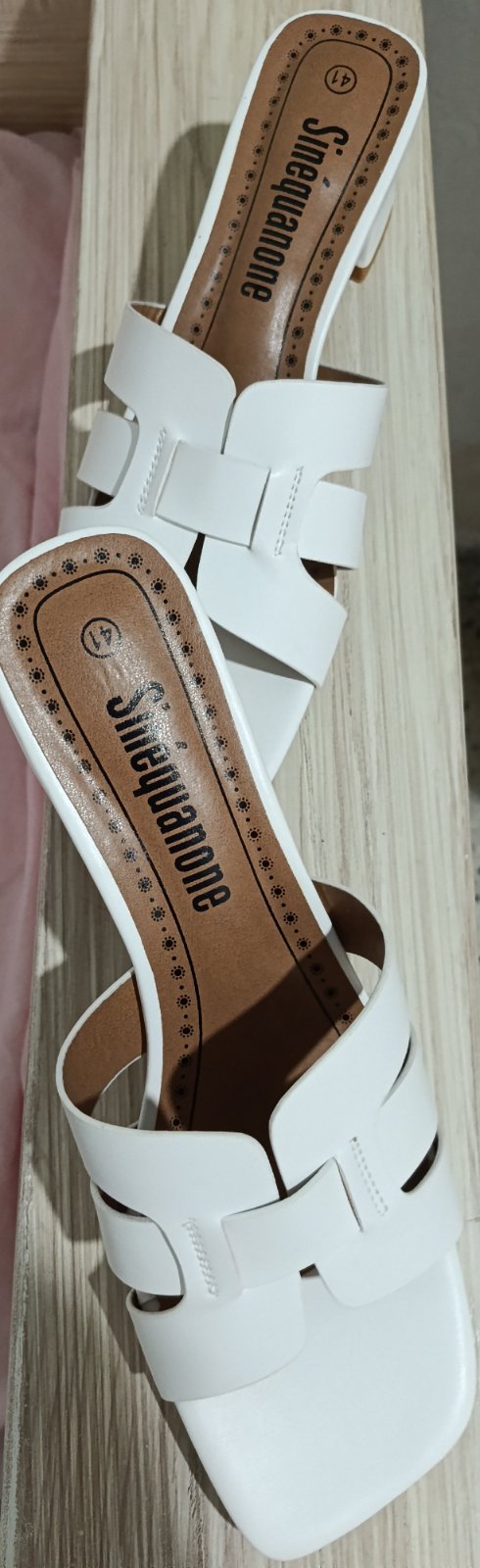 Ce sont des mules blanches à petit talon bloc de la marque Sinéquanone, caractérisées par leurs larges brides entrelacées et leur bout carré