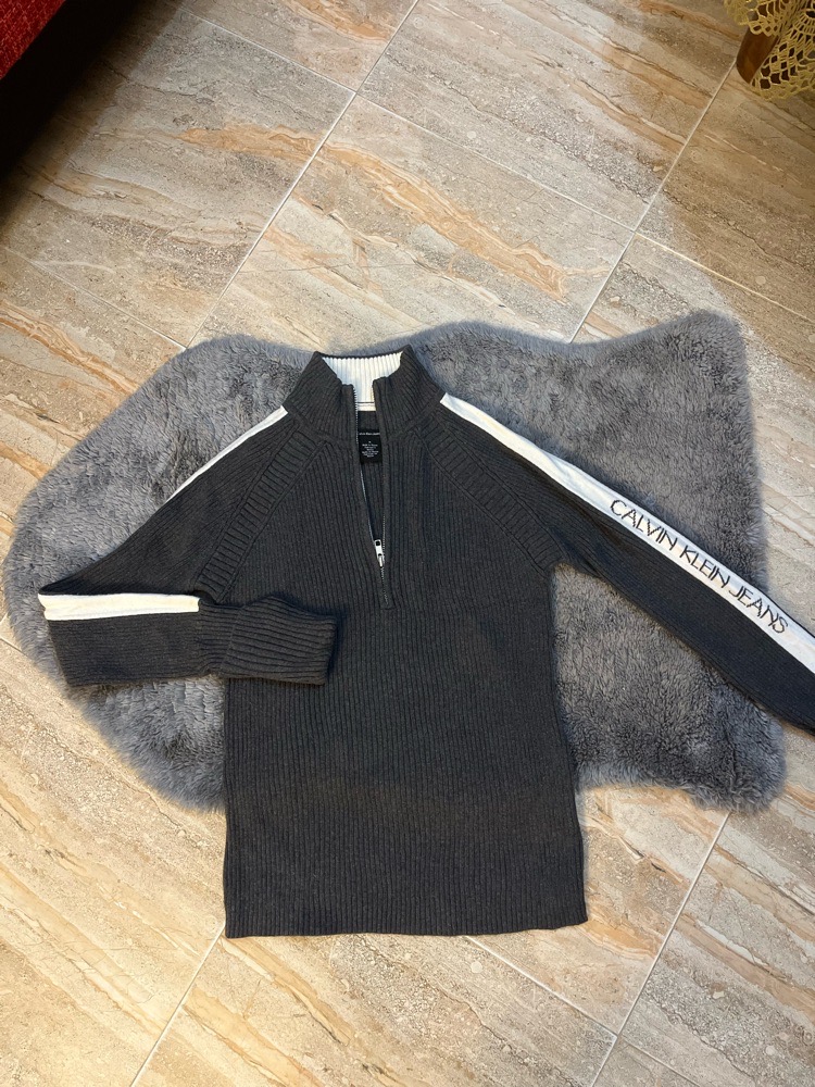 Pull en maille gris foncé à fermeture éclair partielle de Calvin Klein Jeans, avec des bandes blanches sur les manches et le logo de la marque sur la manche gauche