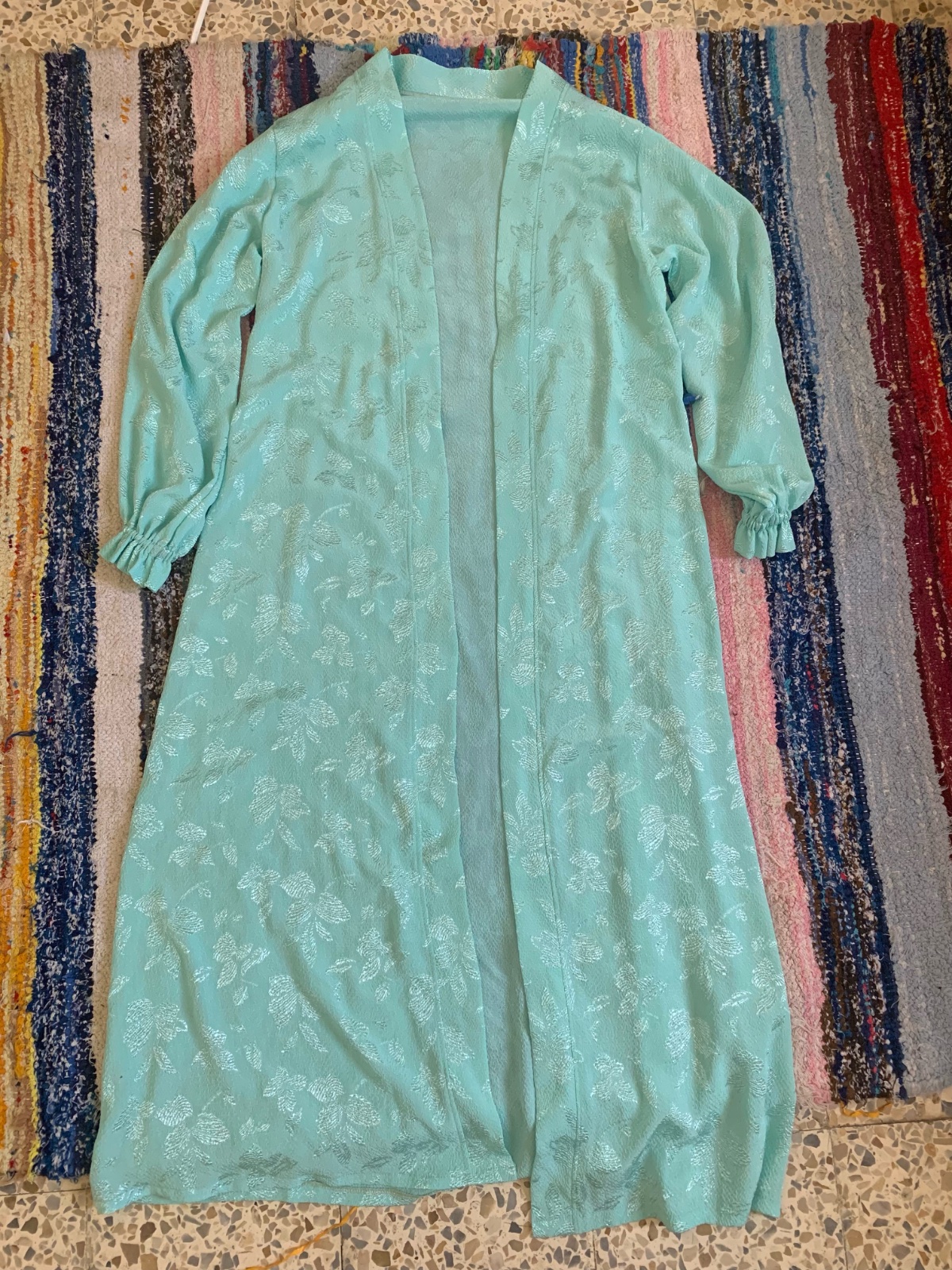 Un long kimono ou un gilet ouvert de couleur bleu menthe, orné de motifs floraux ton sur ton et de manches longues froncées aux poignets.