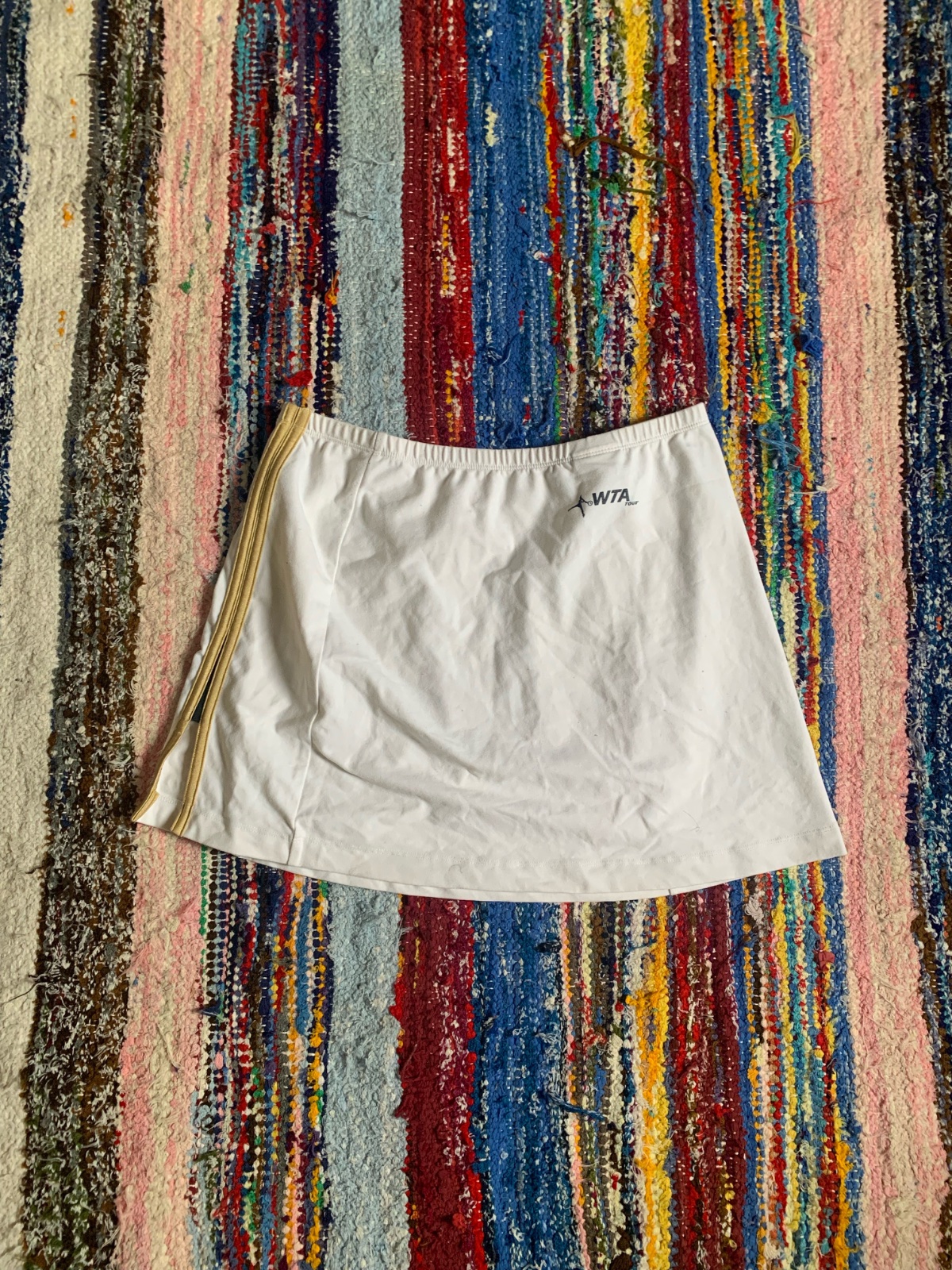 Jupe-short de tennis blanche avec des bandes latérales dorées et le logo WTA Tour imprimé sur la hanche.