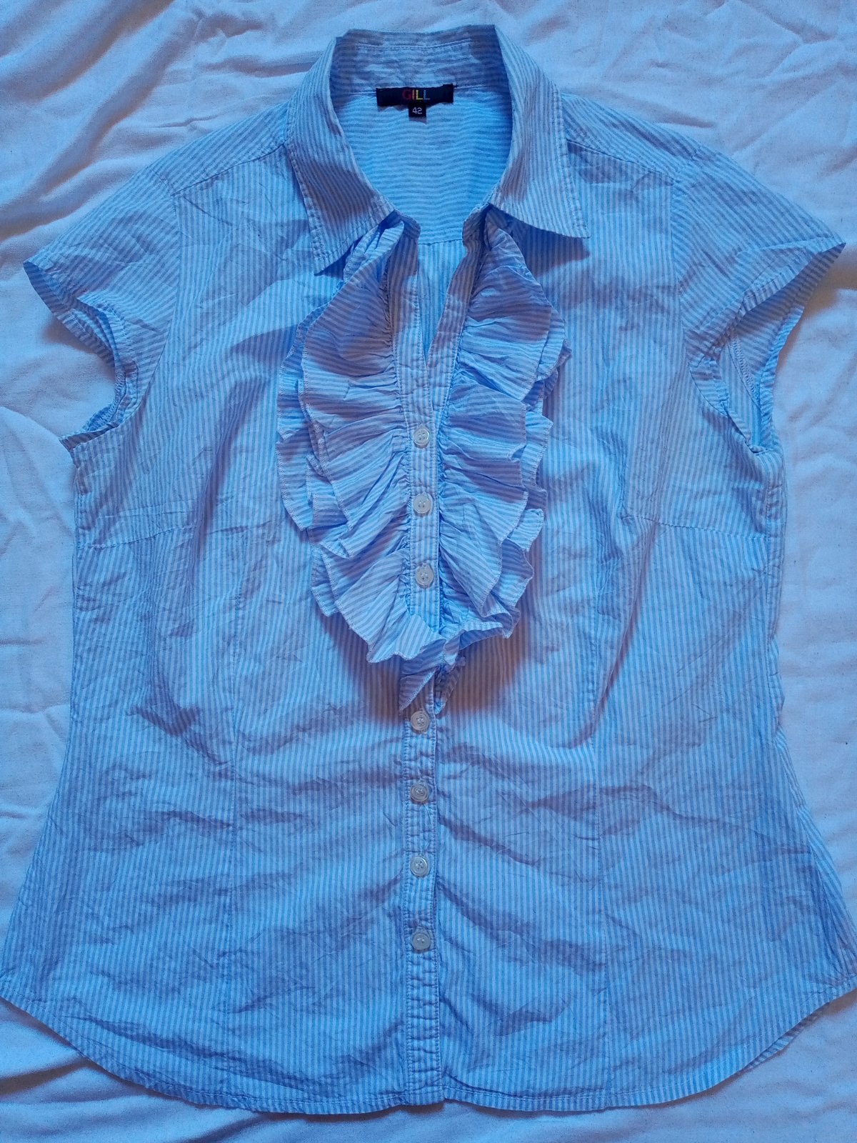 Jolie chemise à manches courtes GILL (taille 42) en excellent état. Elle se distingue par ses fines rayures bleues et blanches et ses magnifiques volants sur le devant qui apportent une touche chic et originale