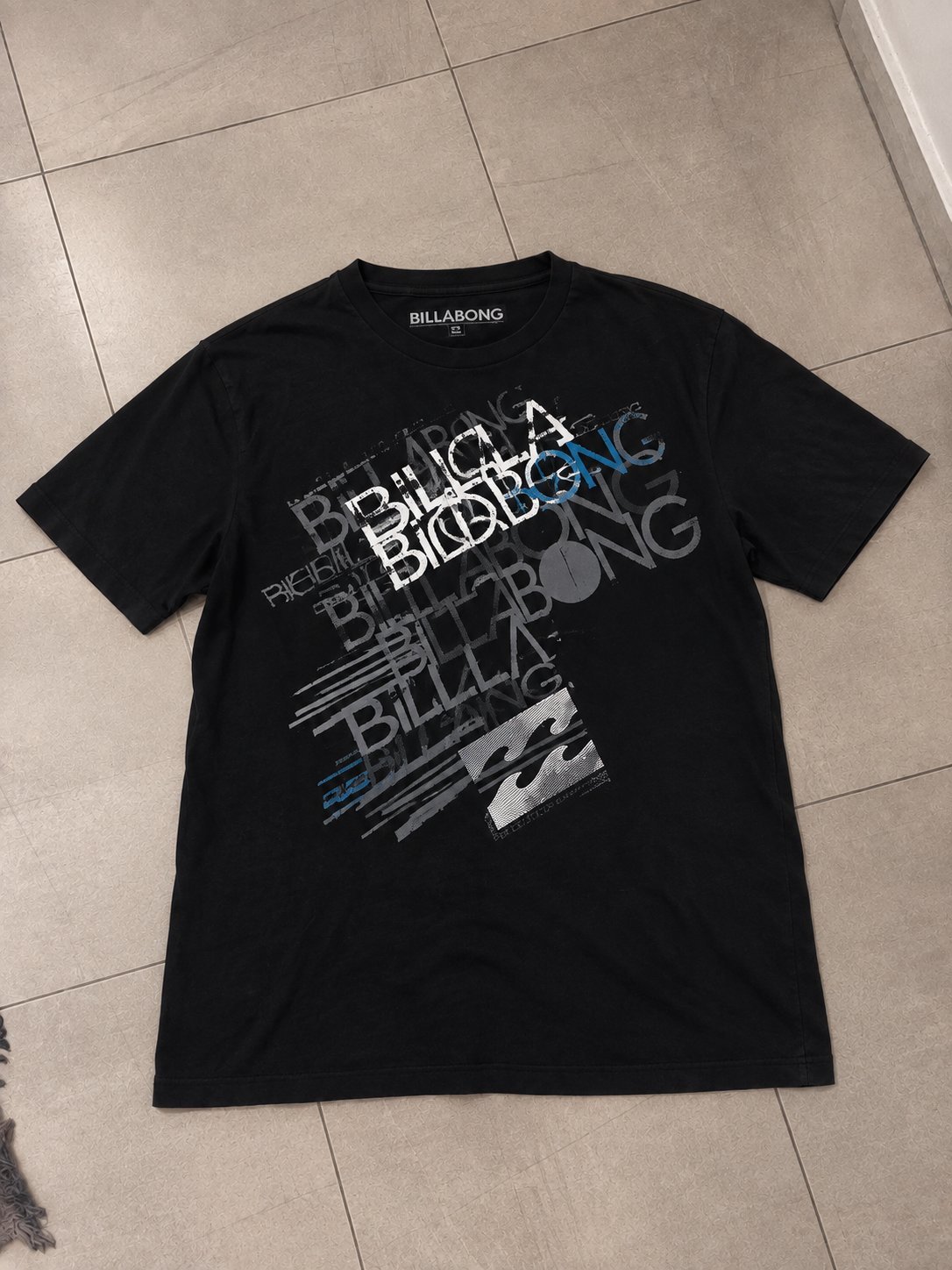 T-shirt Billabong noir, style décontracté, avec un imprimé graphique gris, blanc et bleu.