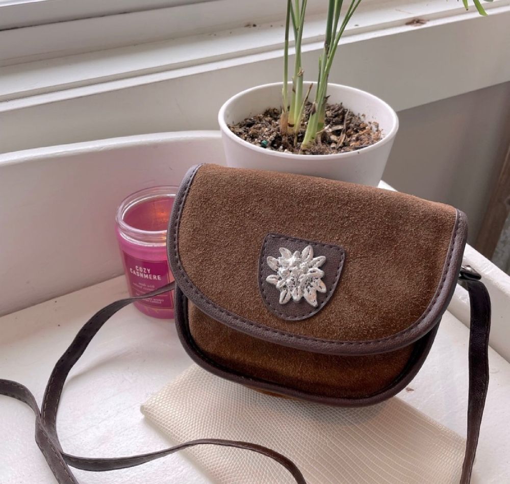C'est un sac à bandoulière de style Trachten en suède vert olive avec des bordures en cuir marron et un insigne floral brodé. 