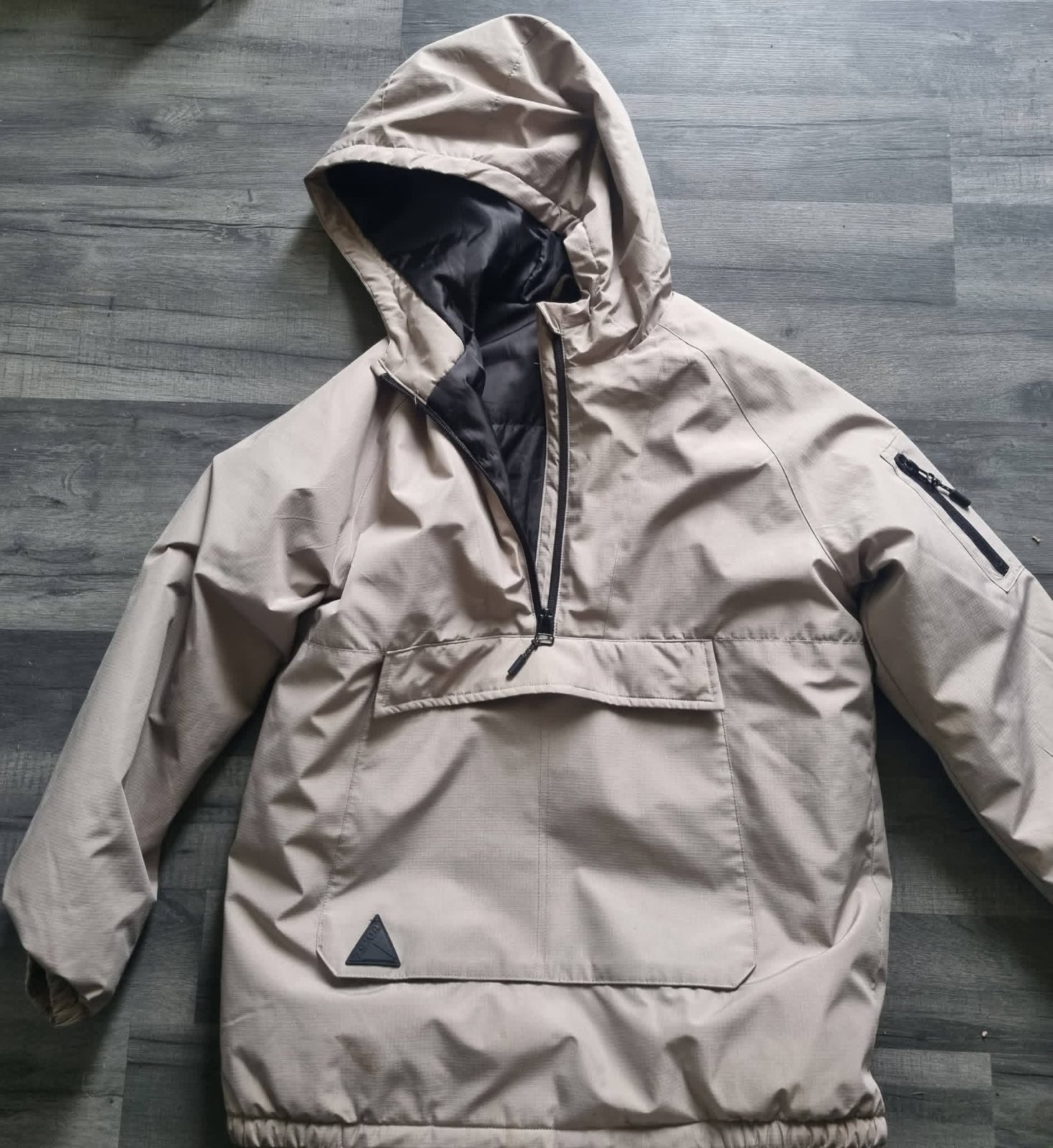 Capuche de couleur beige/taupe, doté d'une fermeture éclair à demi-zip et d'une grande poche centrale à rabat.