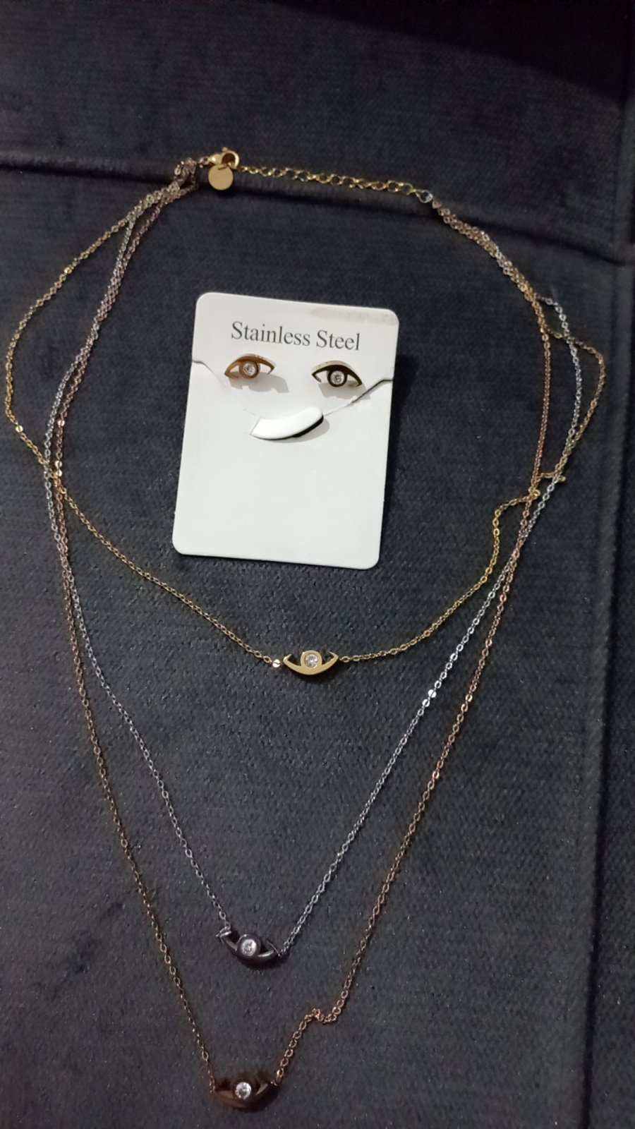 Collier multi-rangs en acier inoxydable se distingue par ses trois chaînes superposées de couleurs différentes (doré, argenté et doré rose) ornées de délicats motifs protecteurs en forme d'œil sertis d'un petit oxyde de zirconium central.