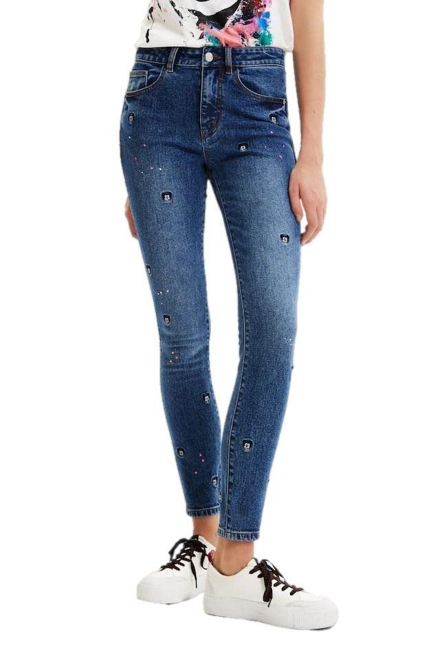 Un jean pour femme orné de strass et de rivets décoratifs, présenté sur un cintre.