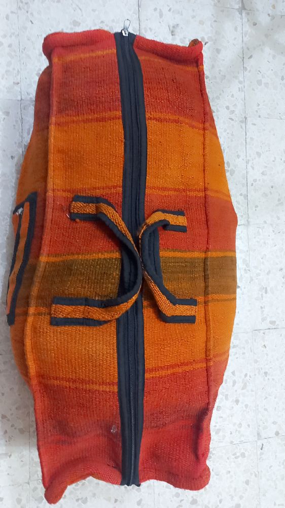 Sac de voyage en laine tissée, avec des motifs à rayures de couleurs orange, rouge et marron. Il est doté d'une fermeture éclair noire et de bretelles noires. Il est difficile de déterminer l'origine exacte ou la marque du sac à dos.