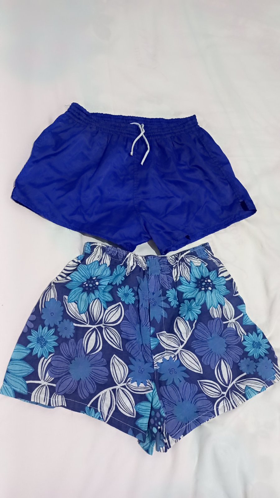 Deux shorts de bain légers se distinguent par leurs nuances de bleu, l'un arborant une teinte unie éclatante avec un cordon de serrage blanc et l'autre un motif floral tropical très estival.
