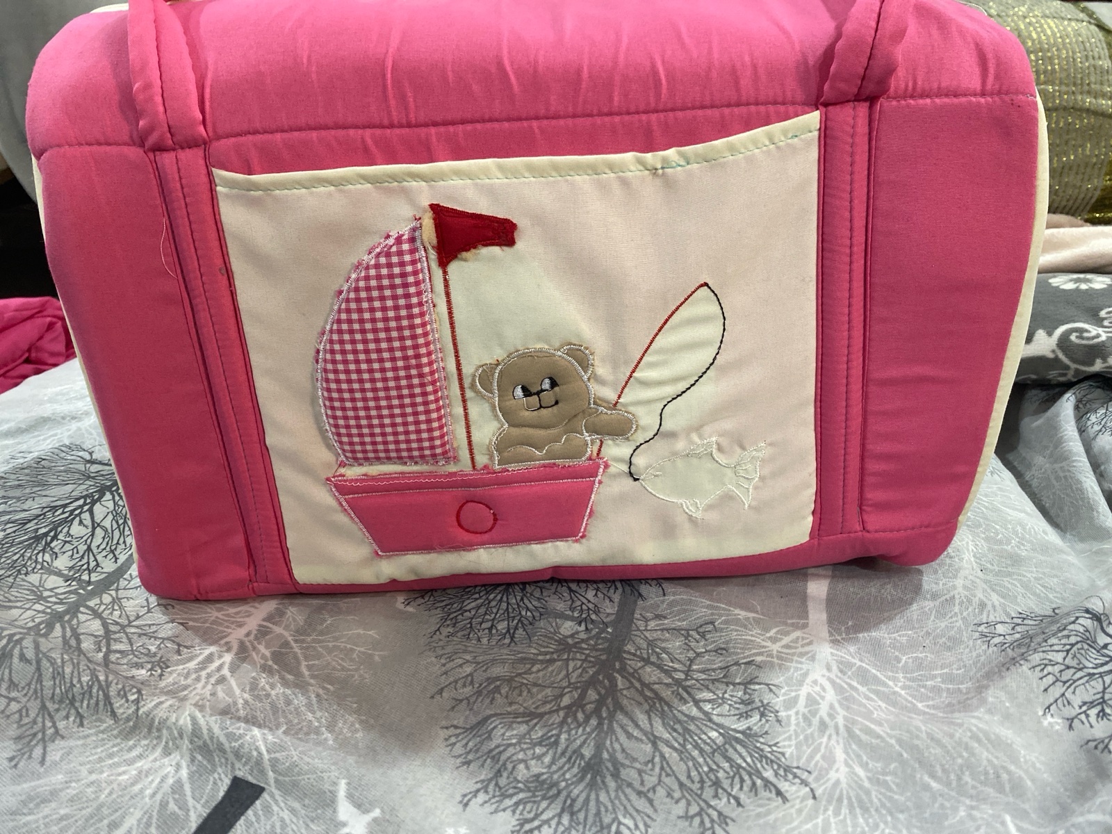Sac à langer (ou couffin nomade) pour bébé, de couleur rose bonbon, doté d'anses de transport et d'un panneau central beige décoré d'une broderie représentant un petit ours brun sous un parasol à carreaux.