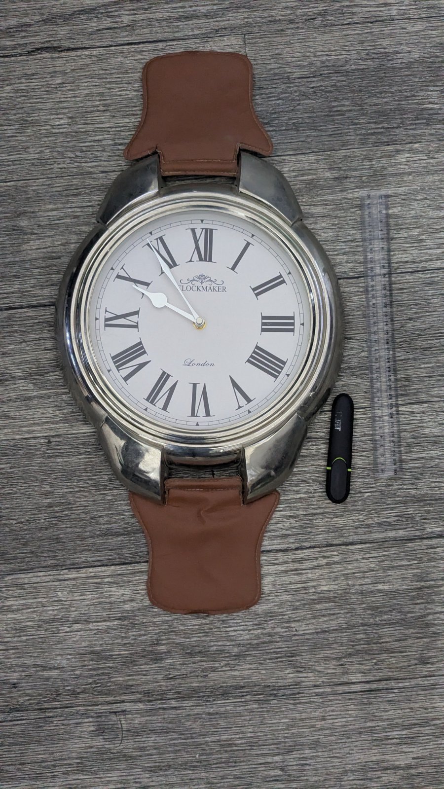 Grande horloge ( en marche)murale décorative conçue pour ressembler à une montre-bracelet surdimensionnée, avec un boîtier en inox, des chiffres romains noirs  et une lanière en cuir marron en haut et en bas.