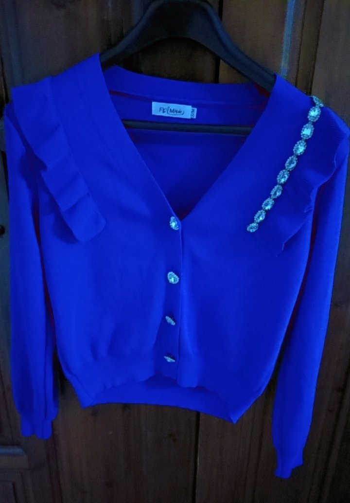 Cardigan bleu  présente une encolure en V, des volants sur les épaules, des boutons brillants et une rangée de strass décoratifs sur l'épaule
