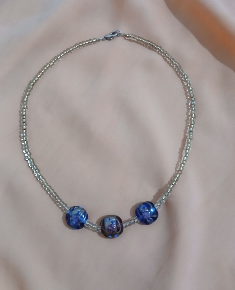 Collier élégant en véritables perles de verre artisanales, monté sur double fil de perles transparentes. Motifs floraux délicats, tons bleus et violet. Fermoir métallique. Pièce faite main, unique.