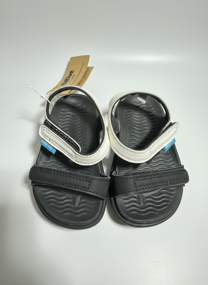 Ces sandales pour enfants sont des Native modèle Charley Sugarlite, caractérisées par leur structure légère en EVA noir avec des sangles contrastantes blanc cassé