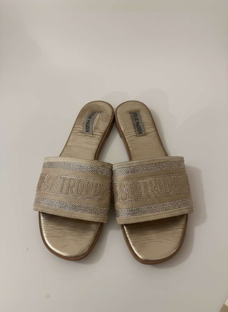 Ces sandales Steve Madden modèle Knox en or se distinguent par une large bride ornée de strass et brodée du mot "ST TROPEZ