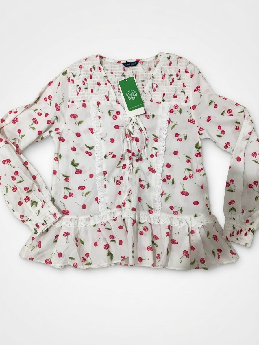 Cette blouse blanche à manches longues se distingue par son charmant imprimé de cerises rouges, son col en V lacé et ses délicats détails de volants en dentelle.