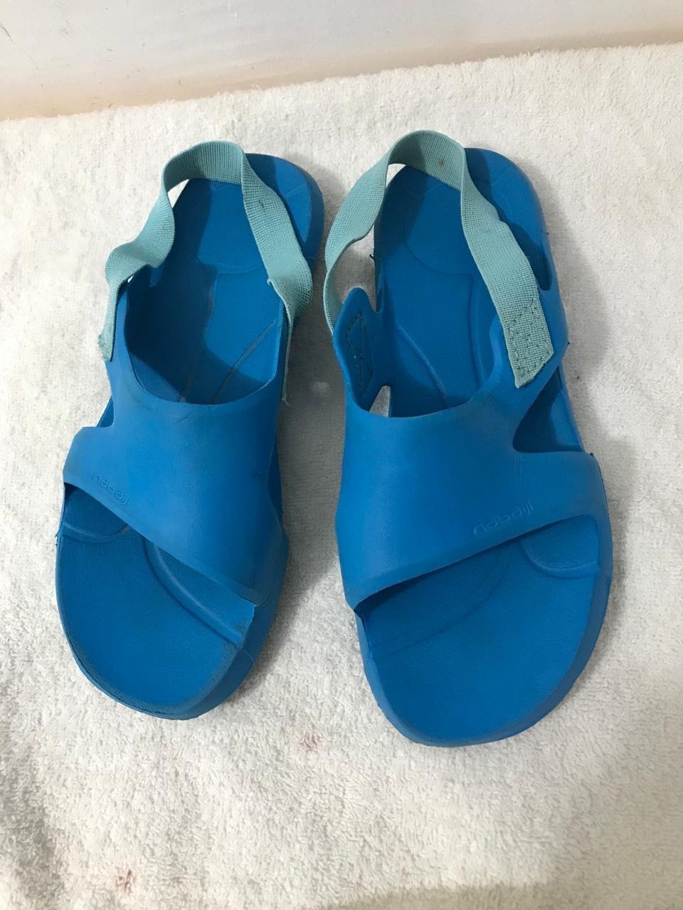 Ces sandales de piscine bleues de la marque Nabaiji (chez [Decathlon]) possèdent une bride élastique turquoise à l'arrière pour un bon maintien du pied.