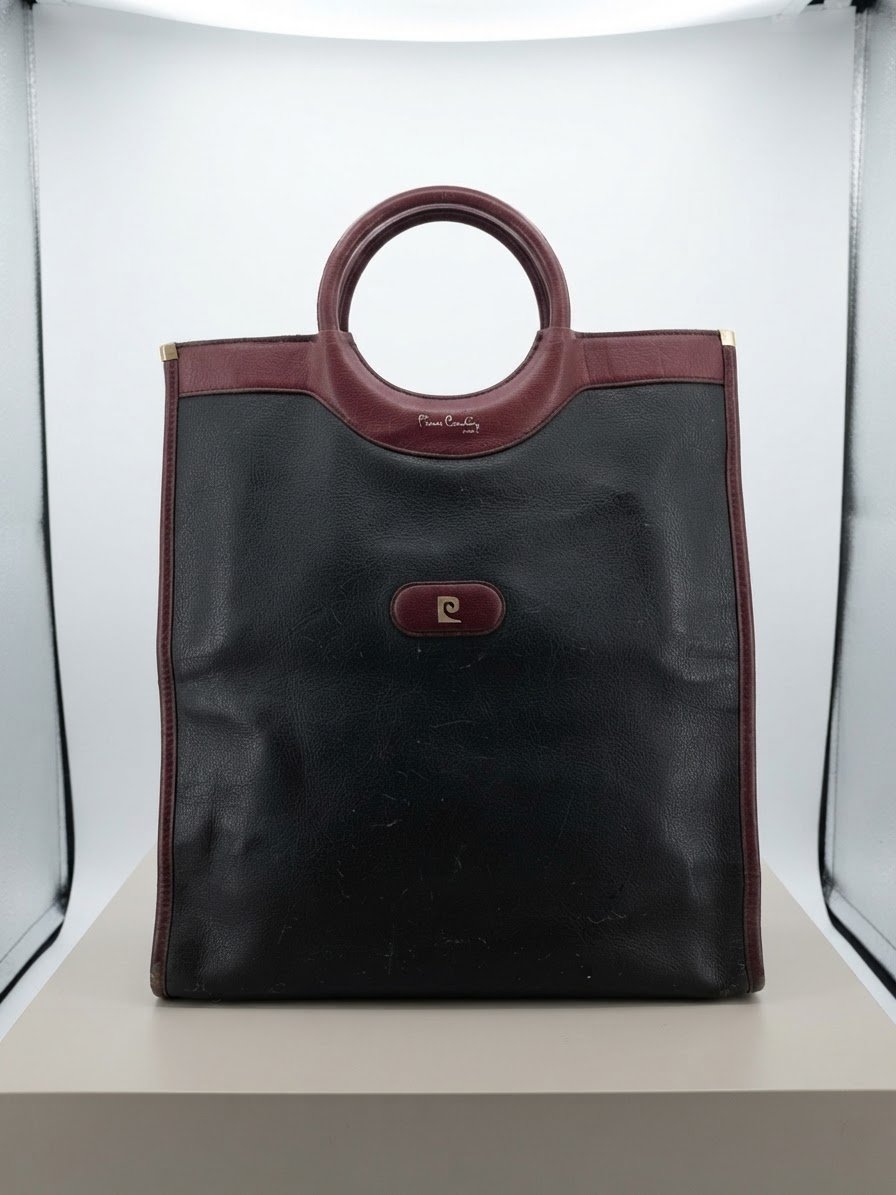 Ce sac cabas vintage Pierre Cardin noir et bordeaux présente des poignées rondes découpées distinctives et le logo métallique de la marque.