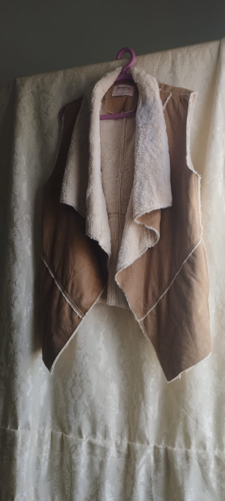Ce gilet sans manches de style mouton retourné est de la marque Bershka, conçu en faux daim marron avec une doublure en fausse fourrure beige. 