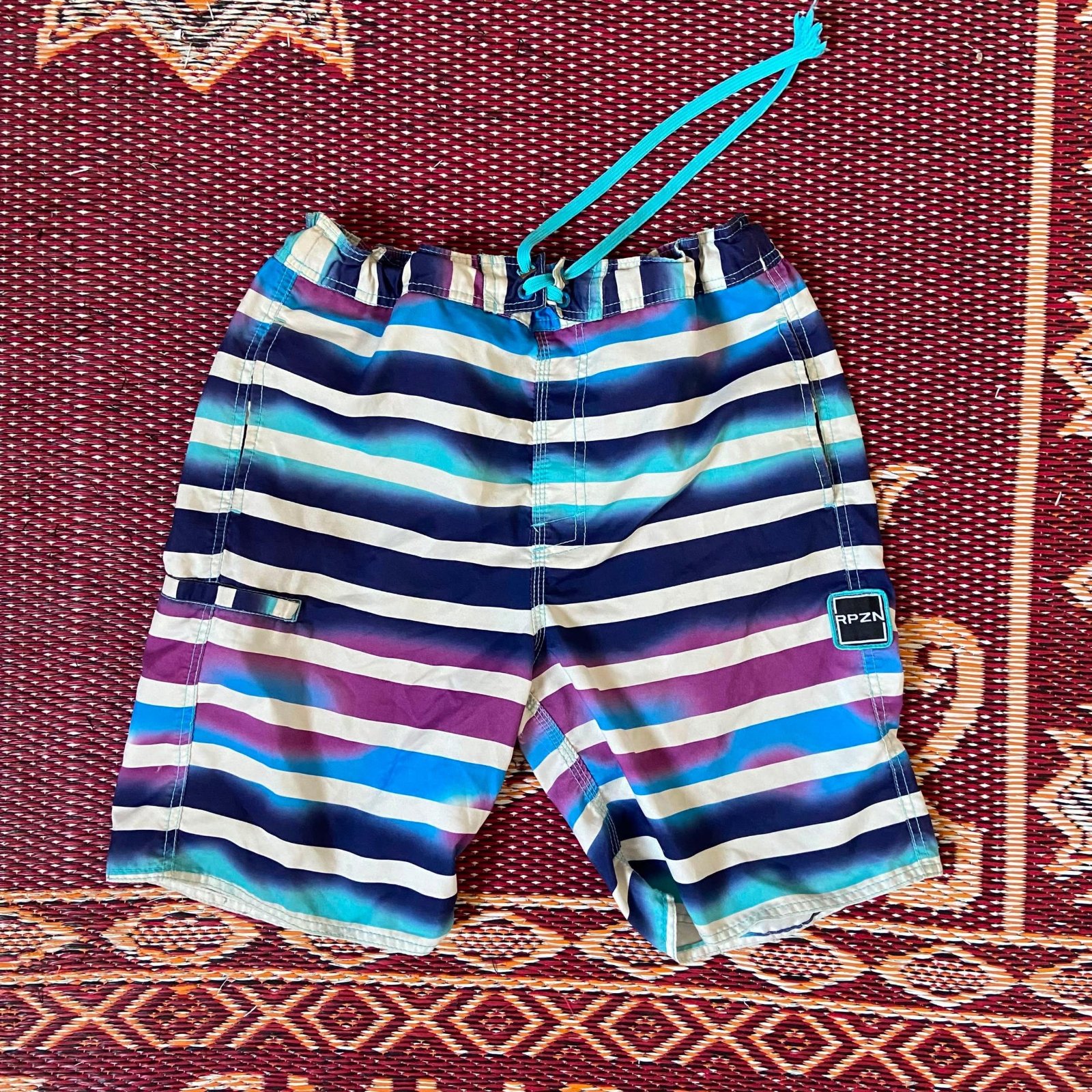 Short de bain rayé bleu, blanc et violet ressemble à un modèle de la marque Kanu Surf et arbore un logo "RPZN" sur la jambe.